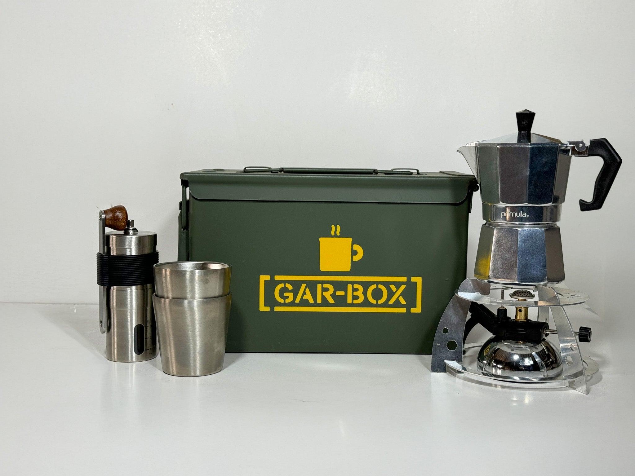 Gar-Box Portable Coffee Bar Box with contents of what comes inside placed in front of the box, items include 2 insulated stainless steel mugs, a portable espresso and coffee maker, a hand-crank coffee grinder, and a mini tabletop butane burner