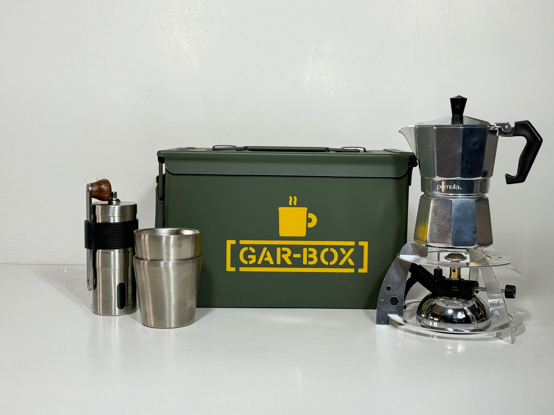 Gar-Box Portable Coffee Bar Box with contents of what comes inside placed in front of the box, items include 2 insulated stainless steel mugs, a portable espresso and coffee maker, a hand-crank coffee grinder, and a mini tabletop butane burner